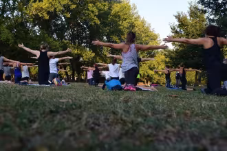 Menschen im Freibad, die sich auf Yoga-Platten im Gras im Freibad mit den H&auml;nden auf dem Boden und den Armen nach links und rechts ausstrecken