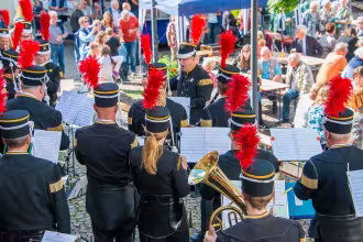 Die im Bild sichtbare Band spielt im Innenraum eines Geb&auml;udes. Im Hintergrund sind Menschen zu sehen, die sich um eine Tafel versammelt haben. Die Bandmitglieder