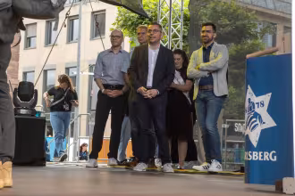 Ein Bild zeigt f&uuml;nf Personen auf einer B&uuml;hne vor einem Geb&auml;ude. Die B&uuml;hne ist mit einem blauen Banner mit einem wei&szlig;en Stern dekoriert.