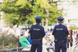 Zwei Polizisten im Hintergrund von einer Stra&szlig;enansammlung mit Menschen. Im Hintergrund ist ein Baum mit gr&uuml;ner Bl&uuml;ten und ein wei&szlig;er Geb&auml;udeteil zu sehen. Im Hintergrund