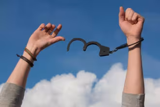 Hand mit Handschuh, an Handgelenk mit Metallkabel und Metallring festgeklemmt, im Hintergrund blauer Himmel mit wenigen wei&szlig;en Wolken.