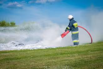Bild: Feuerwehrmann mit Feuerl&ouml;schpumpe in der Hand, um einen Brand zu l&ouml;schen. Im Hintergrund: Wiese mit Gras und B&auml;umen. Im Hintergrund links: Blau-Weiss-Schweigertal-Logo. Im Hintergrund
